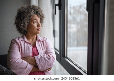 A mature woman of mixed race looking outside a large window, her expression reflecting sadness, concern, and a sense of depression