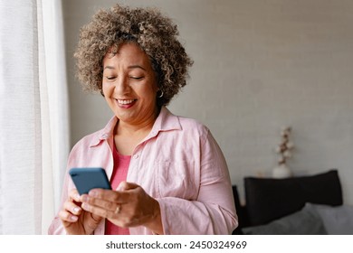 A mature woman of mixed race standing near large window, holding a smartphone and smiling. Mobile phone apps for older people - Powered by Shutterstock