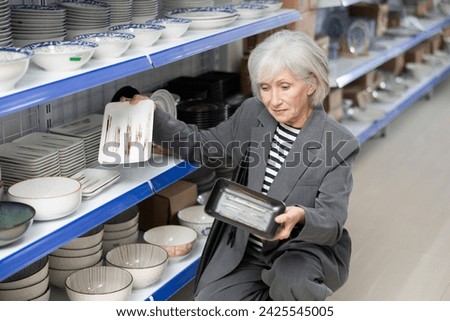 Mature woman near display case with kitchen utensils chooses beautiful clay square plates with pattern. Customer buys view windowshopping and examines ceramic saucers and dishes