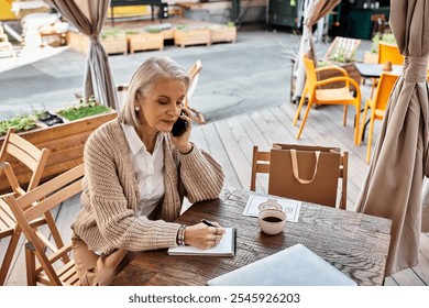 A mature woman is on the phone, taking notes while sipping coffee in a charming cafe. - Powered by Shutterstock