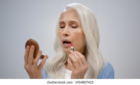 Mature Woman Painting Lips With Lipstick On Isolated Studio Background. Beautiful Old Lady Putting On Makeup Inside. Happy Senior Female Person Looking To Mirror Indoor.