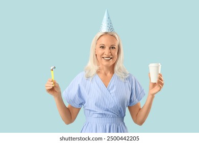 Mature woman in party hat with takeaway cup of coffee and birthday whistle on blue background - Powered by Shutterstock