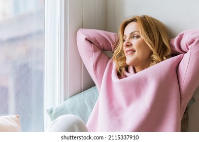 A mature woman in a pink hoodie sits on the windowsill with her arms folded behind her head and enjoys the moment. Slow life concept and digital detox - Shutterstock ID 2115337910