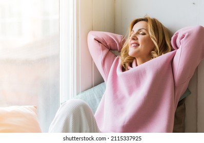 A mature woman in a pink hoodie sits on the windowsill with her arms folded behind her head and enjoys the moment. Slow life concept and digital detox - Shutterstock ID 2115337925