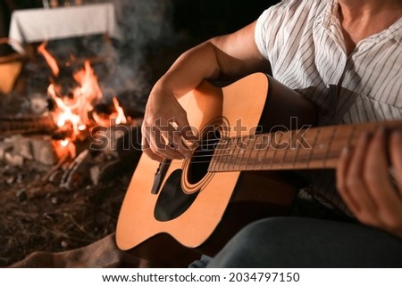 Mature woman playing guitar near fire at barbecue party on summer evening