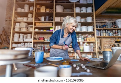 Mature Woman Pottery Artist Using Laptop In Art Studio 
