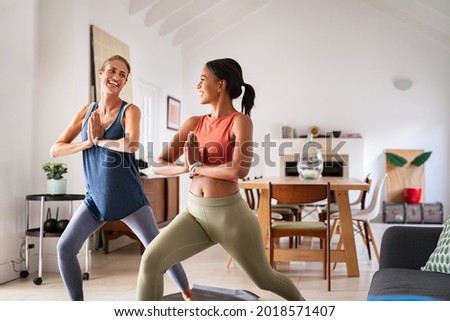 Mature woman practicing yoga at home with mixed race friend. Beautiful sporty indian woman with female friend in yoga position of the warrior at home. Two middle aged lady exercising at home, lockdown