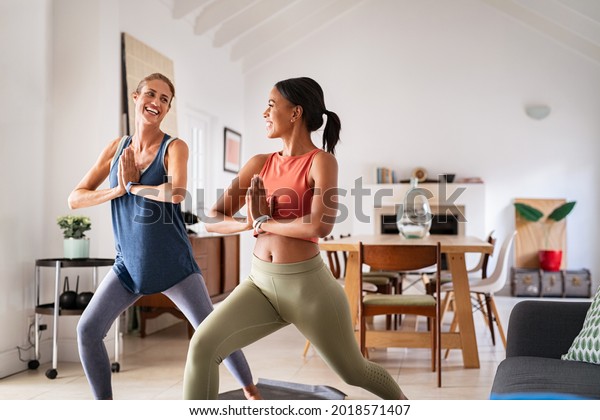 Mature woman practicing yoga at home with mixed\
race friend. Beautiful sporty indian woman with female friend in\
yoga position of the warrior at home. Two middle aged lady\
exercising at home,\
lockdown