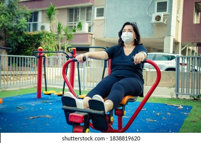 Mature Woman In A Protective Mask Is Strengthening Her Upper Back Muscles On A Rowing Machine. Despite The Pandemic She Works Out Everyday In An Outdoor Fitness Circuit Near Her Home.