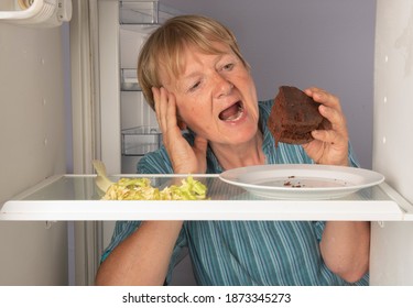 Mature Woman Reaching For Chocolate Cake Inside Fridge Instead Of Vegetables