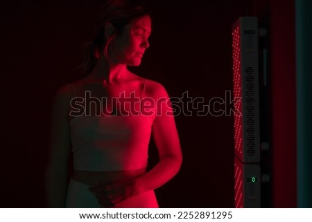 Mature woman receiving red light therapy. Woman standing next to a red light device in a beauty spa. Anti-aging and skin care treatment.