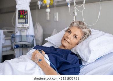 Mature woman recovering from illness while lying on hospital bed with iv drip in hand. Portrait of old hospitalized patient recovering after surgery. Happy senior woman lying on bed in hospital ward. - Shutterstock ID 2151833751