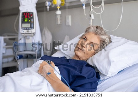 Mature woman recovering from illness while lying on hospital bed with iv drip in hand. Portrait of old hospitalized patient recovering after surgery. Happy senior woman lying on bed in hospital ward.