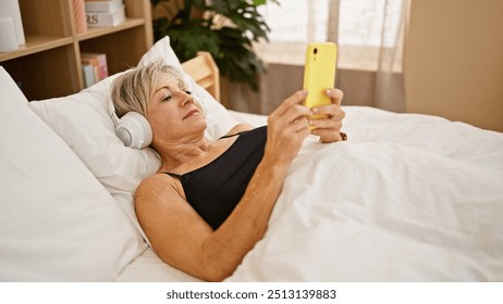 A mature woman relaxes in bed with headphones and a smartphone in a cozy bedroom setting. - Powered by Shutterstock