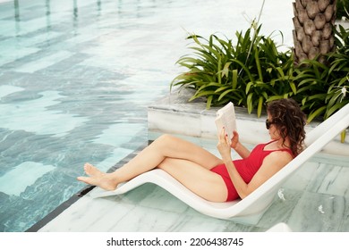 Mature Woman Relaxing On Chaise-lounge By Swimming Pool And Reading Book