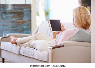 Mature Woman Relaxing On Sofa At Home Using Digital Tablet