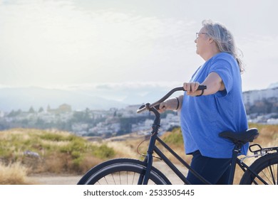 A mature woman rides a black retro-style bicycle against a cloudy mountain backdrop with a village in the background - Powered by Shutterstock