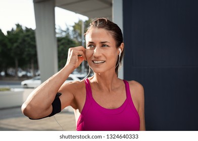 Mature Woman Runner Wearing Armband And Listening To Music On Earphones. Fit Sportswoman Taking A Break From Outdoors Training And Looking Away. Portrait Of Smiling Woman Running On City Street.