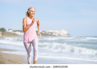 Mature Woman Running Along The Shore Of The Beach. Older Female Doing Sport To Keep Fit