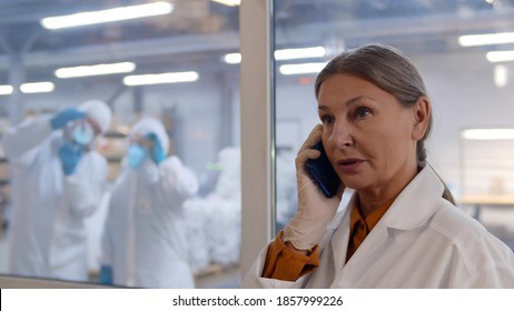 Mature Woman Scientist In White Coat And Gloves Talking On Phone At Modern Chemical Factory. Female Laboratory Worker Having Phone Call With Colleagues In Overalls Carrying Experiment Behind Window