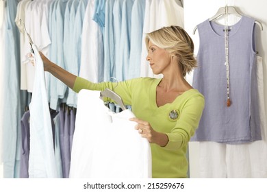 Mature Woman Shopping In Clothes Shop, Comparing Two Tops On Coathangers, Side View