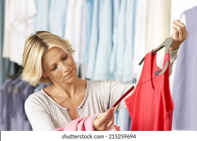 Mature Woman Shopping In Clothes Shop, Holding Red Vest Top On Coathanger, Checking Price Tag, Smiling
