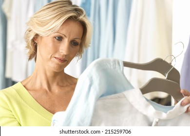 Mature Woman Shopping In Clothes Shop, Comparing Two Tops On Coathangers, Close-up