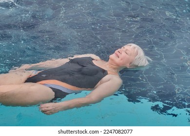 Mature woman with short gray hair, eyes closed, in swimsuit lying on back on water, relaxing. Outdoor thermal pool. Mental health concept - Shutterstock ID 2247082077