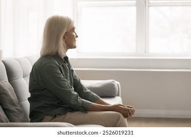 Mature woman sits on couch and gazing out of window, deep in thoughts, reflecting on her physical well-being, managing chronic conditions, or worrying about future health issues. Solitude, retirement - Powered by Shutterstock