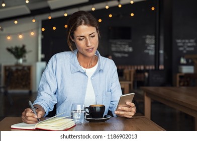 Mature woman sitting in cafe at table and typing a message on smartphone. Middle aged blogger making notes using application. Businesswoman reading from smartphone while working in cafe interior. - Powered by Shutterstock