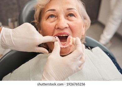 Mature woman sitting at the dentist chair with open mouth. Cropped view of the female dentist putting in artificial teeth to her senior patient mouth in stomatology clinic - Powered by Shutterstock