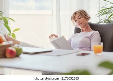 Mature Woman Sitting With Feet Up On The Desk And Reading A Book