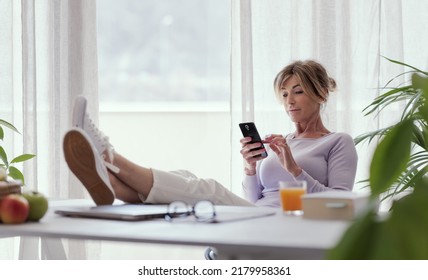 Mature Woman Sitting With Feet Up On The Desk At Home, She Is Connecting With Her Smartphone
