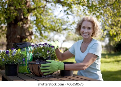 A Mature Woman Sitting At A Garden Bench Planting Hanging Baskets