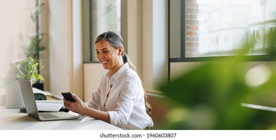 Mature woman sitting at office desk with mobile phone and laptop. Businesswoman in office using phone.