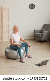 Mature Woman Sitting On The Ball In Front Of Laptop Computer And Exercising In The Room