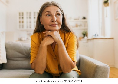 Mature woman sitting on couch putting chin on crossed hands, looking aside with positively thoughtful face expression, waiting to her husband returning from work, isolated on kitchen background - Powered by Shutterstock