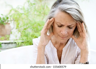 Mature Woman Sitting On A White Sofa In A Home Garden Touching Her Head With Her Hands While Having A Headache Pain And Feeling Unwell, Outdoors.