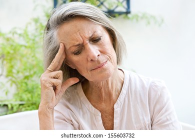 Mature Woman Sitting On A White Sofa In A Home Garden Touching Her Head With Her Hand And Fingers While Having A Headache Pain And Feeling Unwell, Outdoors.