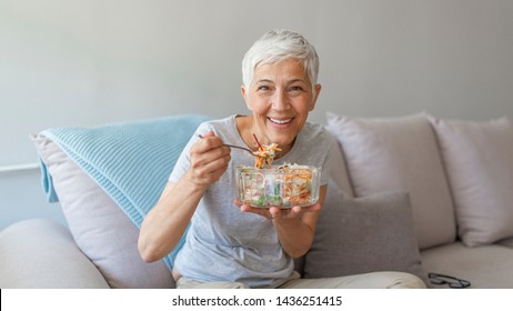 Mature Woman Sitting And Relaxing On A White Couch At Home While Eating A Small Green Salad, Home Interior. Senior Woman Eating Healthy Food. Well Being Indoors. Lifestyle, Smiling.