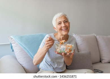 Mature Woman Sitting And Relaxing On A White Couch At Home While Eating A Small Green Salad, Home Interior. Senior Woman Eating Healthy Food. Well Being Indoors. Lifestyle, Smiling.