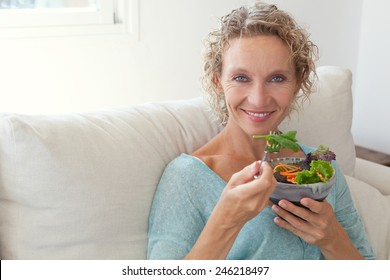 Mature Woman Sitting And Relaxing On A White Couch At Home While Eating A Small Green Salad, Home Interior. Senior Woman Eating Healthy Food. Well Being Indoors. Lifestyle, Smiling.