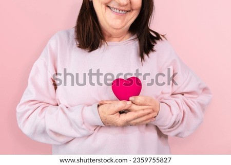  A mature woman smiles while holding a pink resin heart, symbolizing hope and support for breast cancer awareness, ideal for October Pink campaigns