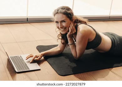 Mature woman smiling at the camera while joining a virtual yoga class on a laptop. Happy woman following an online workout tutorial at home. Cheerful senior woman lying on an exercise mat. - Powered by Shutterstock