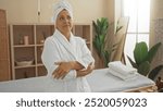 Mature woman standing with arms crossed in a spa room, wearing a white robe and towel on her head, surrounded by wellness decor including plants and massage table