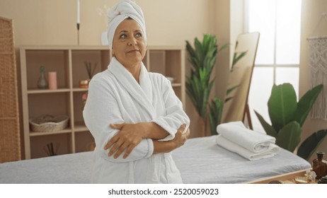 Mature woman standing with arms crossed in a spa room, wearing a white robe and towel on her head, surrounded by wellness decor including plants and massage table - Powered by Shutterstock