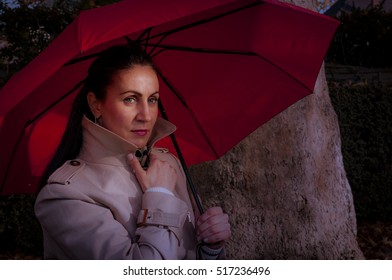 Mature Woman Is Standing Outdoors In A Park Next To A Tree At Night After Sunset Wearing A Trench Coat Holding A Red Umbrella Not Looking At The Camera