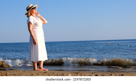 Mature Woman In A Straw Hat And White Dress Enjoying The Sun Walking Along The Blue Sea Coast On A Sunny Summer Day, Enjoying Freedom And Relaxation. The Concept Of A Senior Citizen Everyday Life