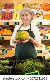 Mature Woman Supermarket Worker Standing In Fruits And Vegetables Department And Holding Melon In Hands.