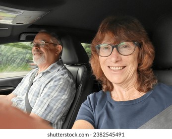 Mature woman takes a selfie while her partner is driving a car. They are both wearing seat belts and smiling as they go on a road trip together. Adventure day out for middle aged couple. - Powered by Shutterstock